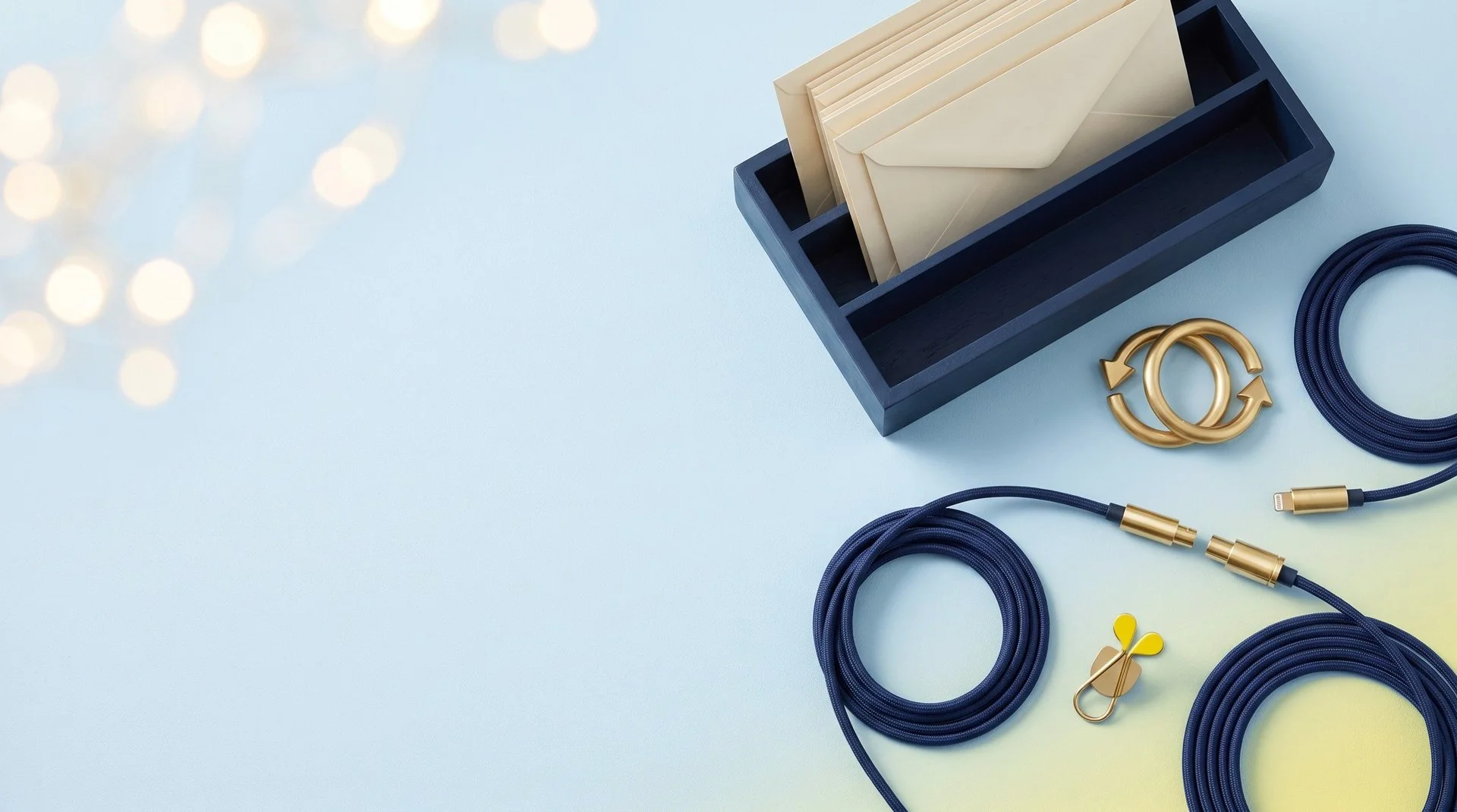 A navy mail sorting tray of cream envelopes, two navy cables with brass connectors about to plug together and a brass sync sculpture on a pale blue backdrop fading to light yellow in the corner.